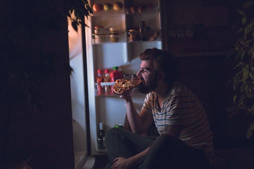 young man eating unheathy food at night sitting infront of the refrigerator